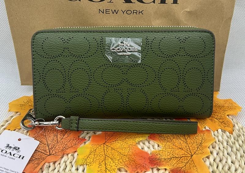 Green Coach long zip-around wallet with perforated signature pattern and wrist strap, shown in front of a Coach shopping bag.