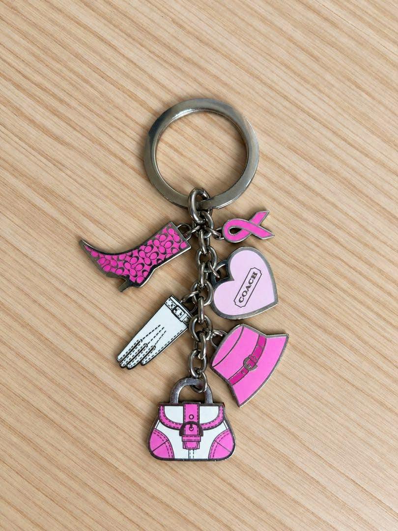 Front view of a COACH pink bag charm keyring with multiple pink charms (handbag, hat, boot, heart, glove, ribbon) on a wood surface.