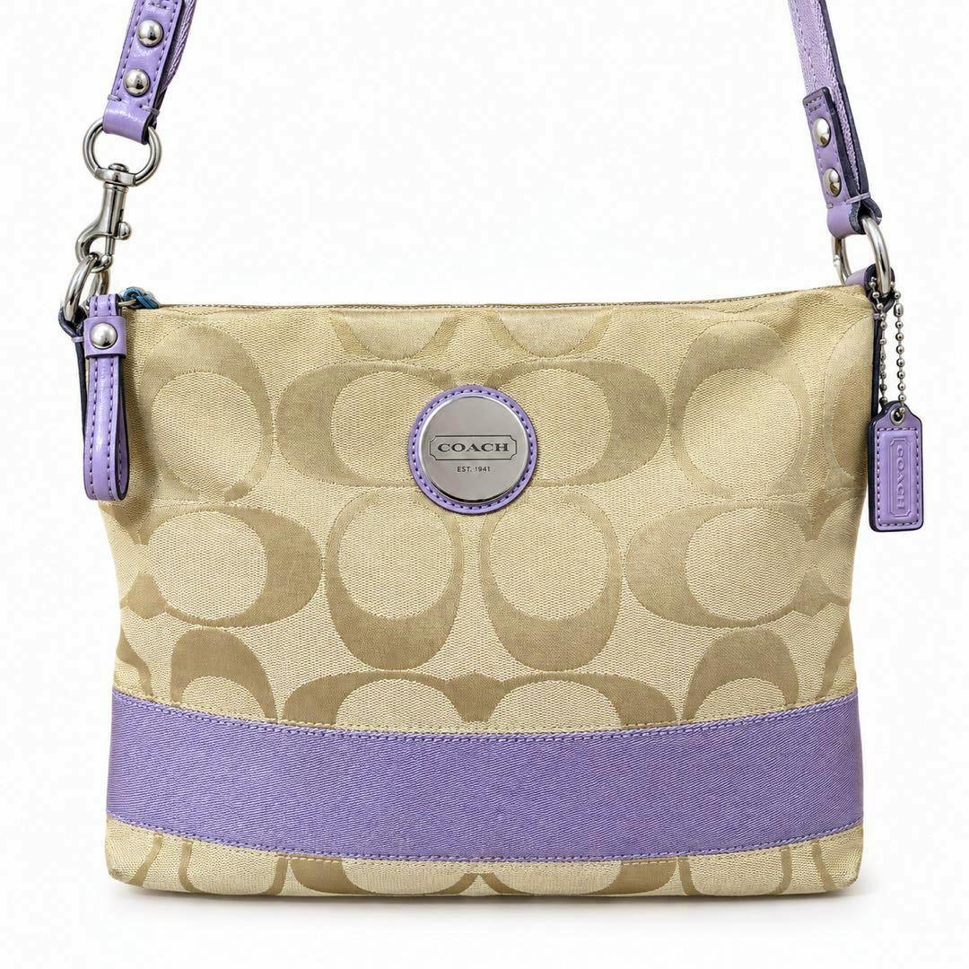 Front view of a COACH signature canvas shoulder crossbody bag in beige with purple trim, strap, and logo medallion.