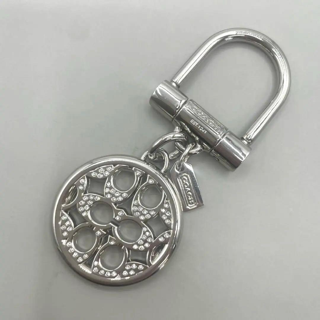 Close-up of a silver Coach signature round rhinestone keychain with a D-ring attachment on a gray background.