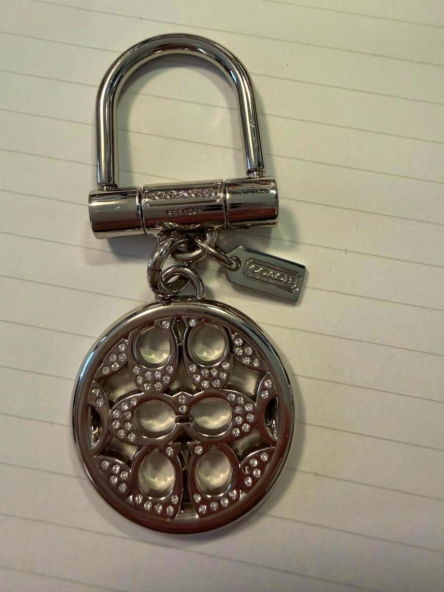 Silver Coach round bag charm with signature pattern and rhinestone accents attached to a silver keyring.