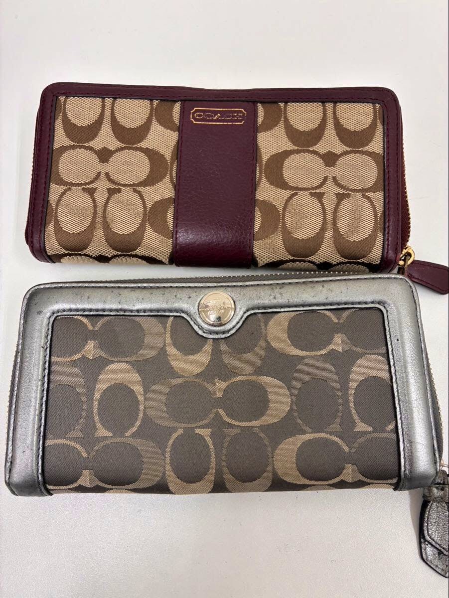 Two Coach signature wallets side by side on a white background, one with brown fabric and burgundy leather trim and the other with brown fabric and metallic silver trim.