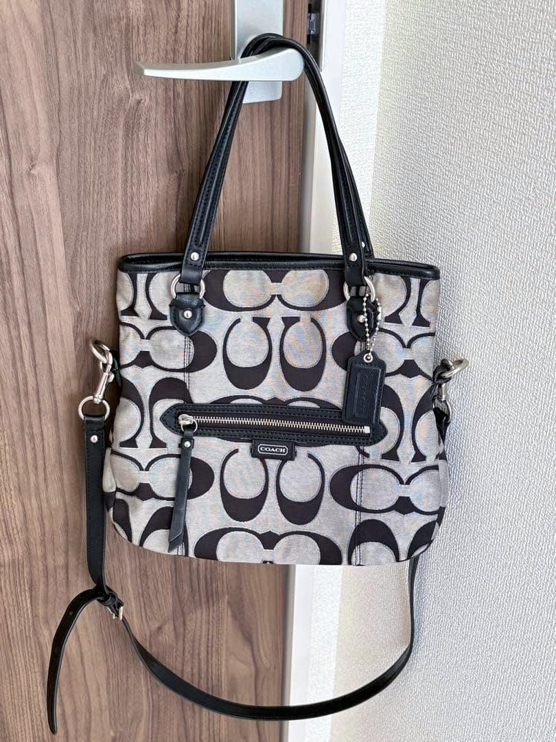 Coach signature satin carryall 2way canvas bag with black 'C' pattern, two top handles, front zip pocket and a detachable shoulder strap hanging on a door handle.