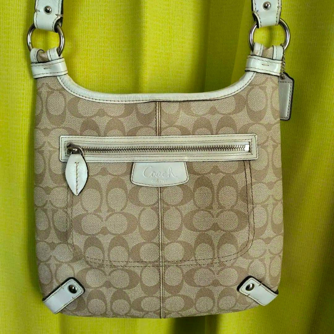 Front view of a beige and ivory COACH signature canvas shoulder bag with a front zip pocket and white leather trim.