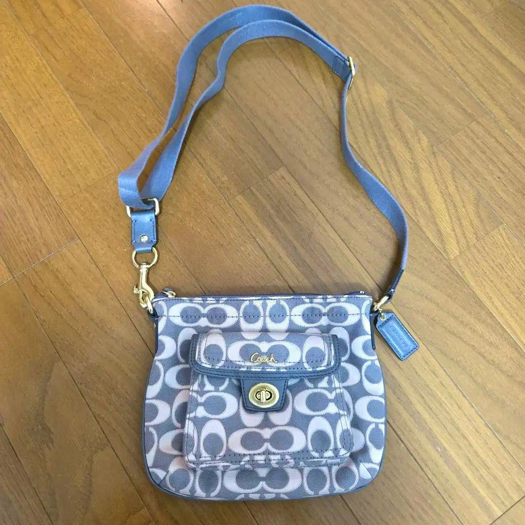 Gray Coach signature canvas shoulder bag with front flap pocket and turn-lock closure, shown with its adjustable strap on a wood floor.