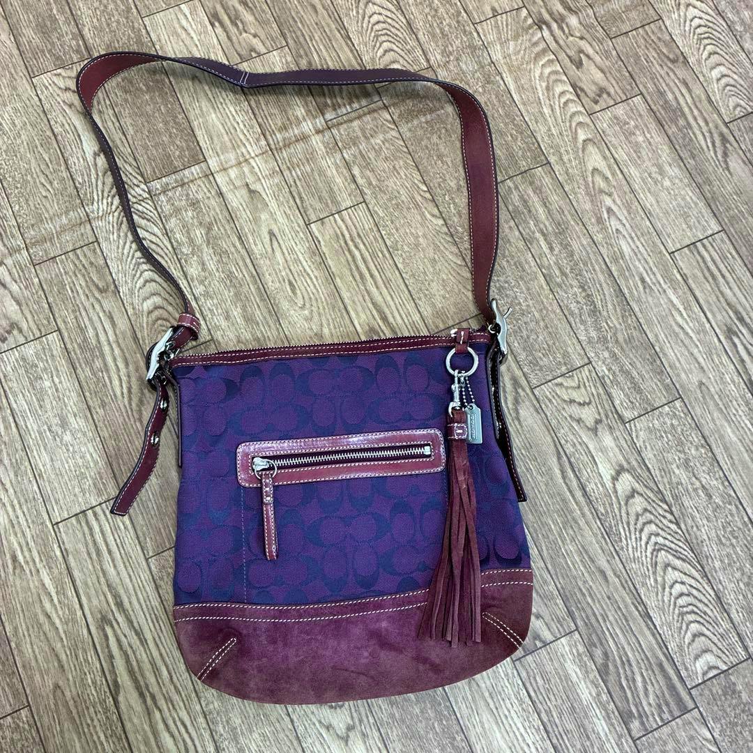 Purple COACH signature canvas shoulder bag with front zip pocket, suede bottom, leather strap, and tassel charm laid on a wood floor.