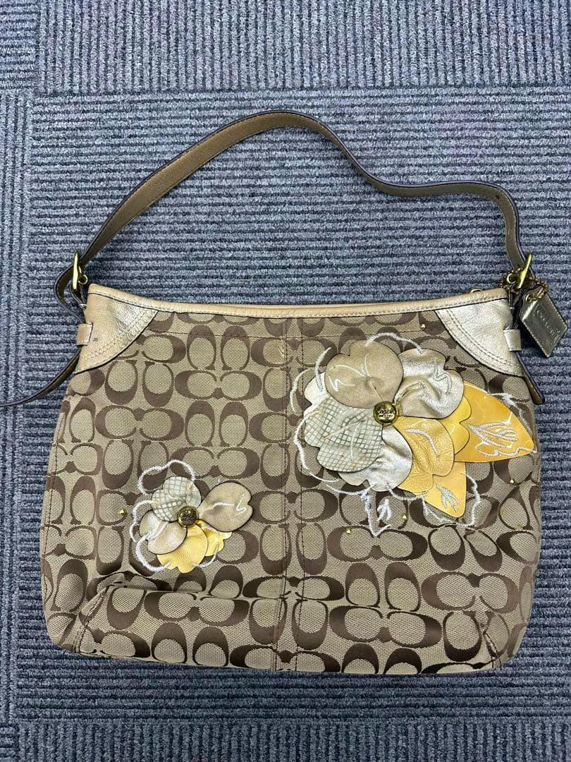 Front view of a Coach signature canvas shoulder bag with floral appliqué and a single shoulder strap lying on a carpet.