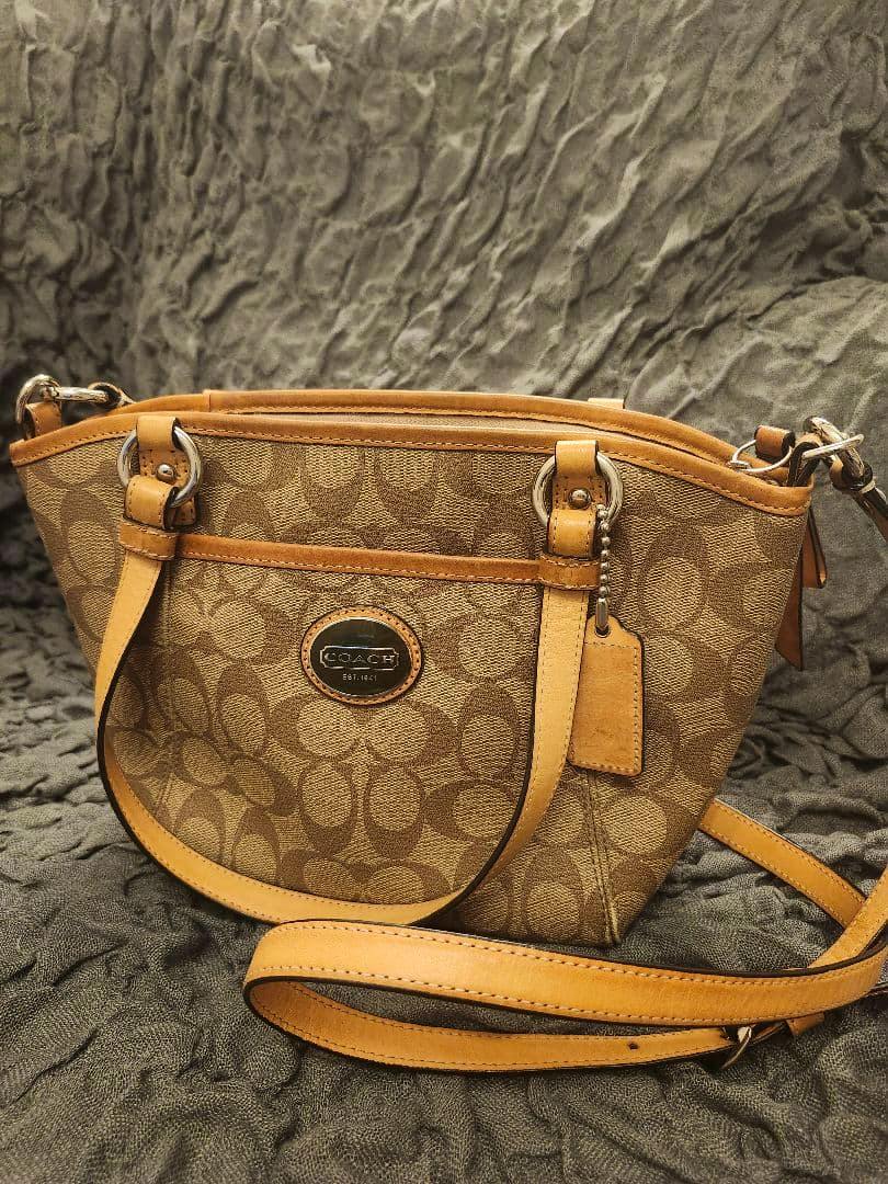 Front view of a brown COACH signature canvas shoulder bag with leather trim, metal logo plaque, and a long shoulder strap on a textured fabric background.