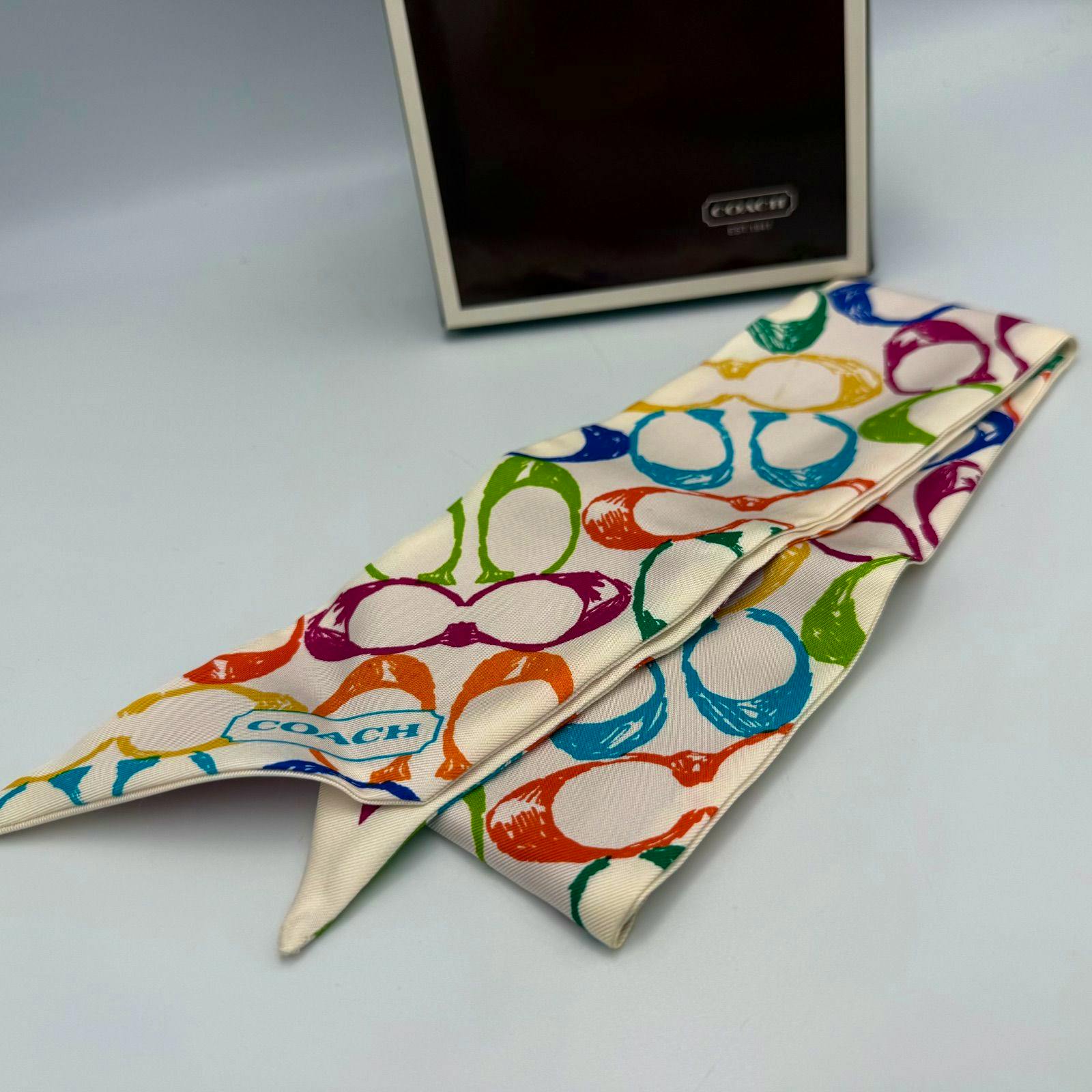 Folded multicolor Coach silk twilly scarf showing the signature pattern with a Coach box in the background.