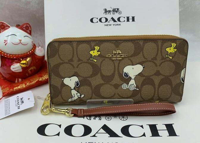 Coach signature brown zip-around long wallet featuring Snoopy and Woodstock prints with a brown wrist strap, shown in front of a Coach box.