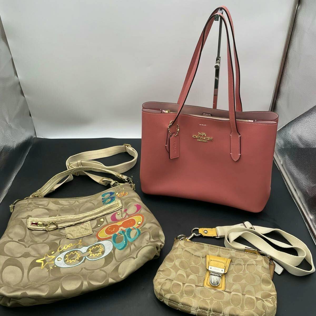 Three COACH bags: a pink tote and two tan signature canvas bags displayed on a black surface.