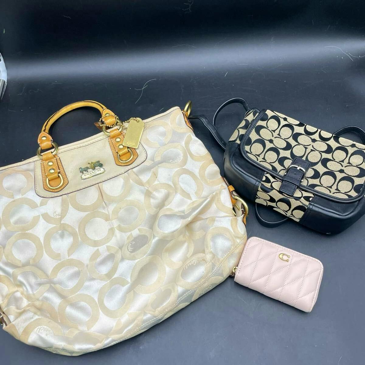 Three Coach items on a dark background: a large cream signature tote, a black signature shoulder bag, and a small pink quilted mini wallet.