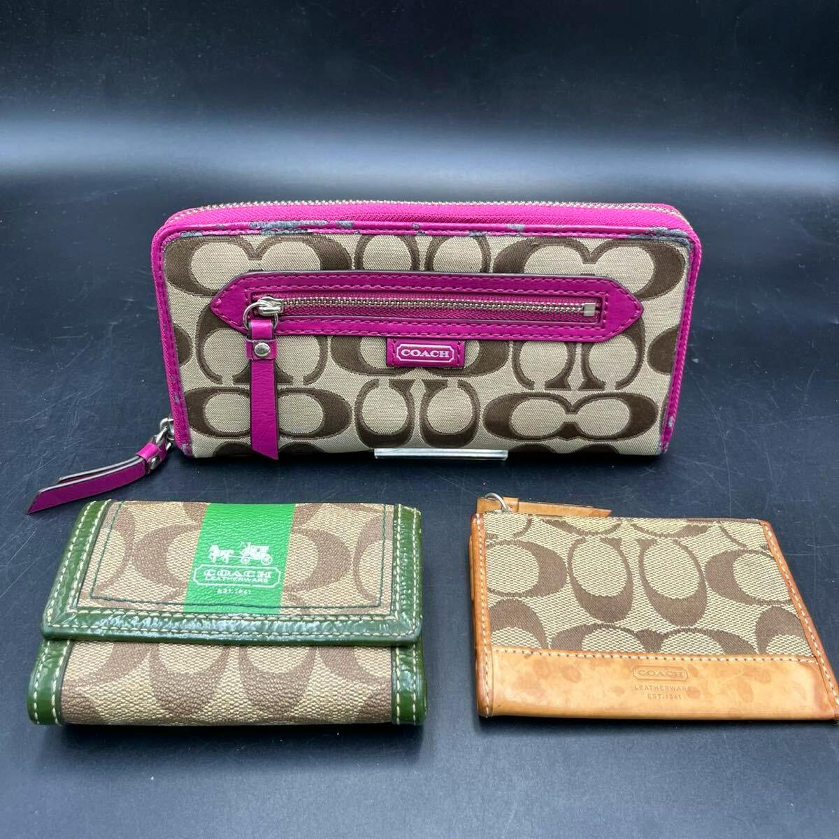 Three Coach canvas wallets on a dark background: a large zippered signature canvas wallet with pink trim behind two smaller signature canvas wallets with green and tan trims.