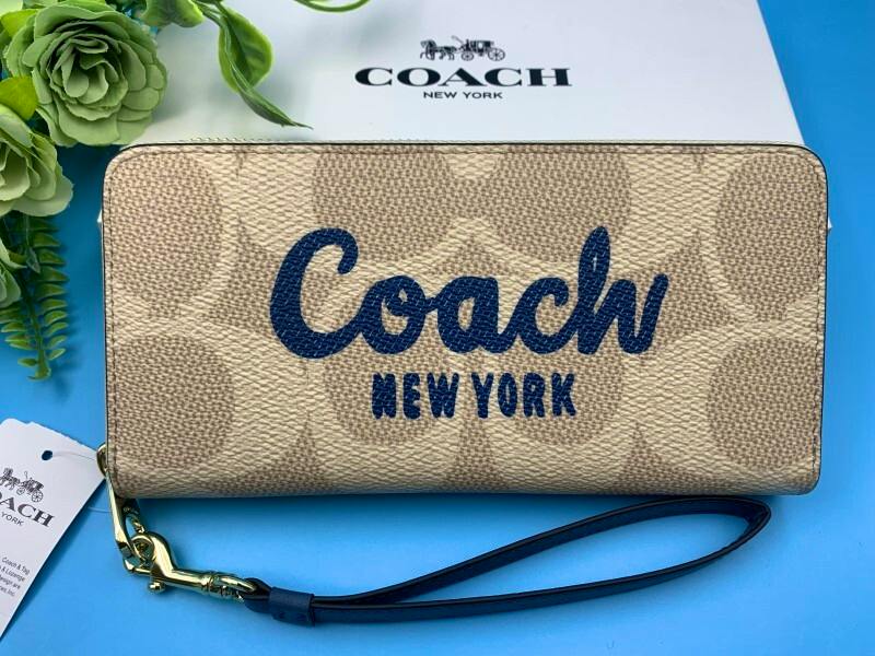 Beige COACH signature long zip-around wallet in cow leather with wrist strap and tag displayed on a blue background.