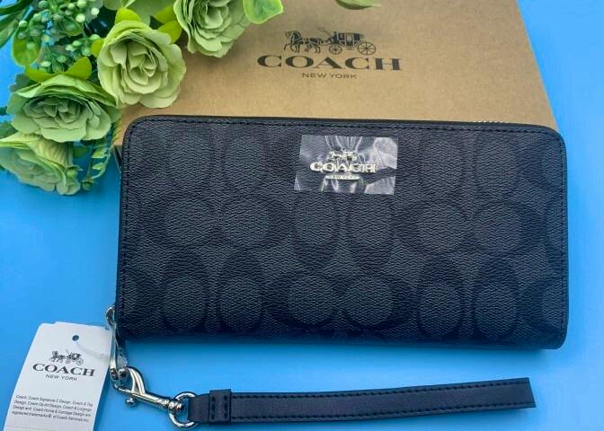 COACH long zip-around wallet in dark signature print shown front-facing with a detachable wrist strap and branded box behind.