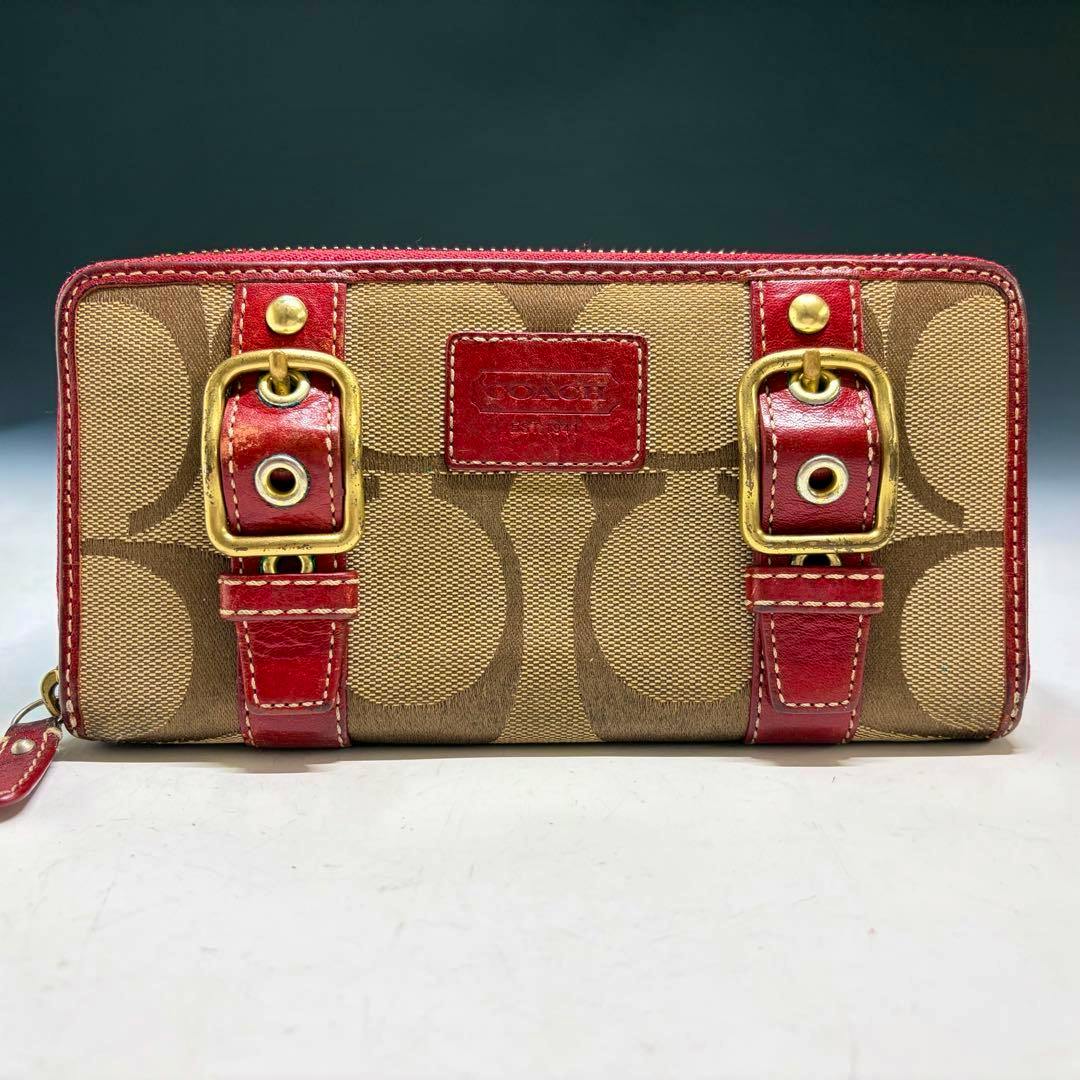Front view of a COACH signature canvas long zip-around wallet showing beige signature print, red leather trim, gold buckles, and a red logo patch.