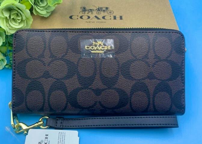 Brown COACH signature long zip-around cow leather wallet with gold logo and wrist strap on a blue surface, with a COACH box and tag visible.