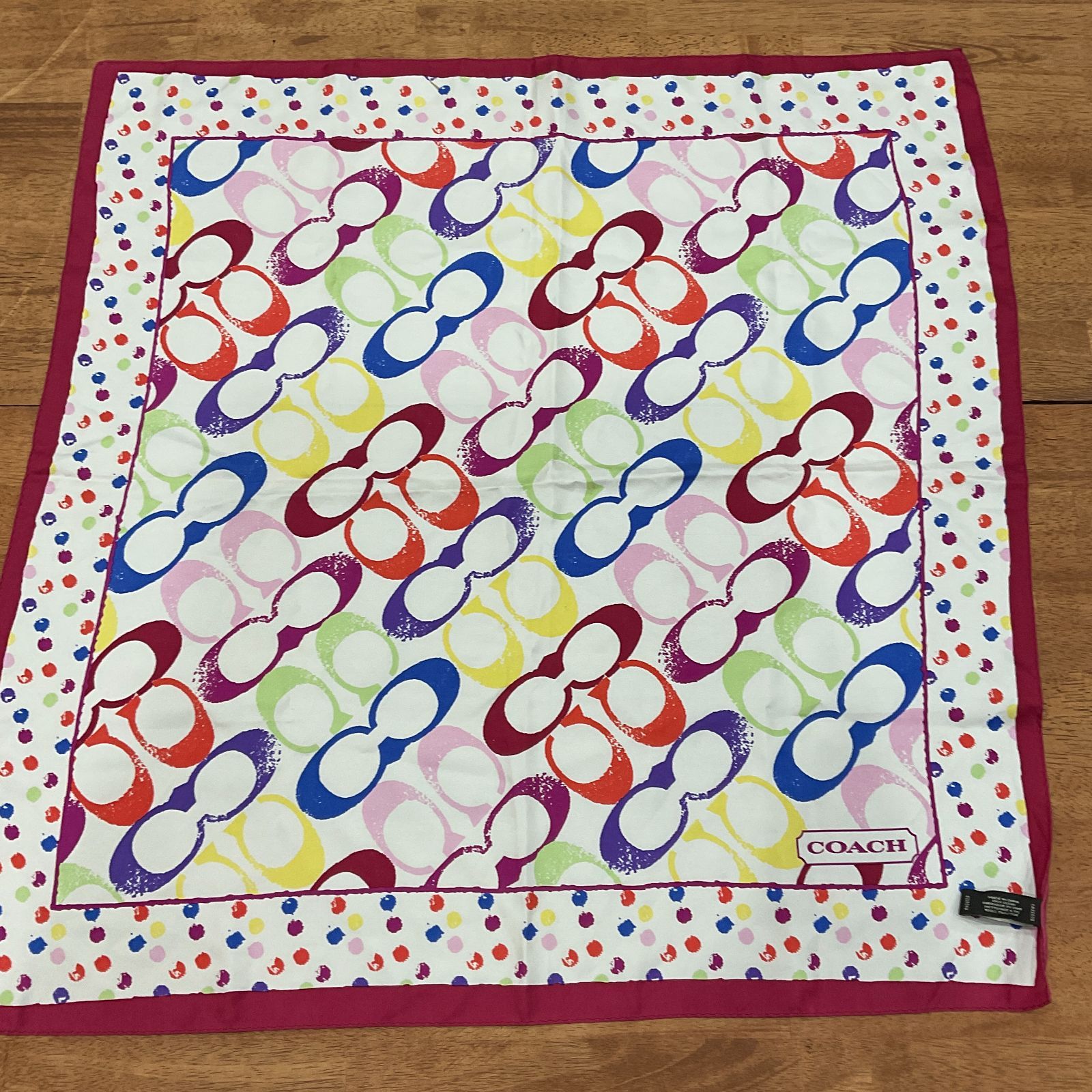 Laid flat Coach silk scarf with diagonal multicolored linked‑C pattern, polka‑dot border, and visible Coach tag on a wooden floor.