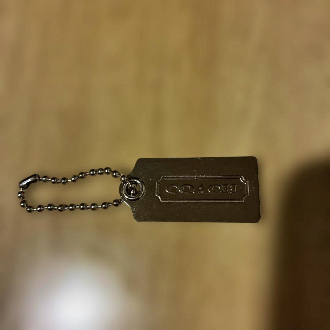 Silver rectangular metal COACH keychain tag with attached ball chain on a wooden surface.