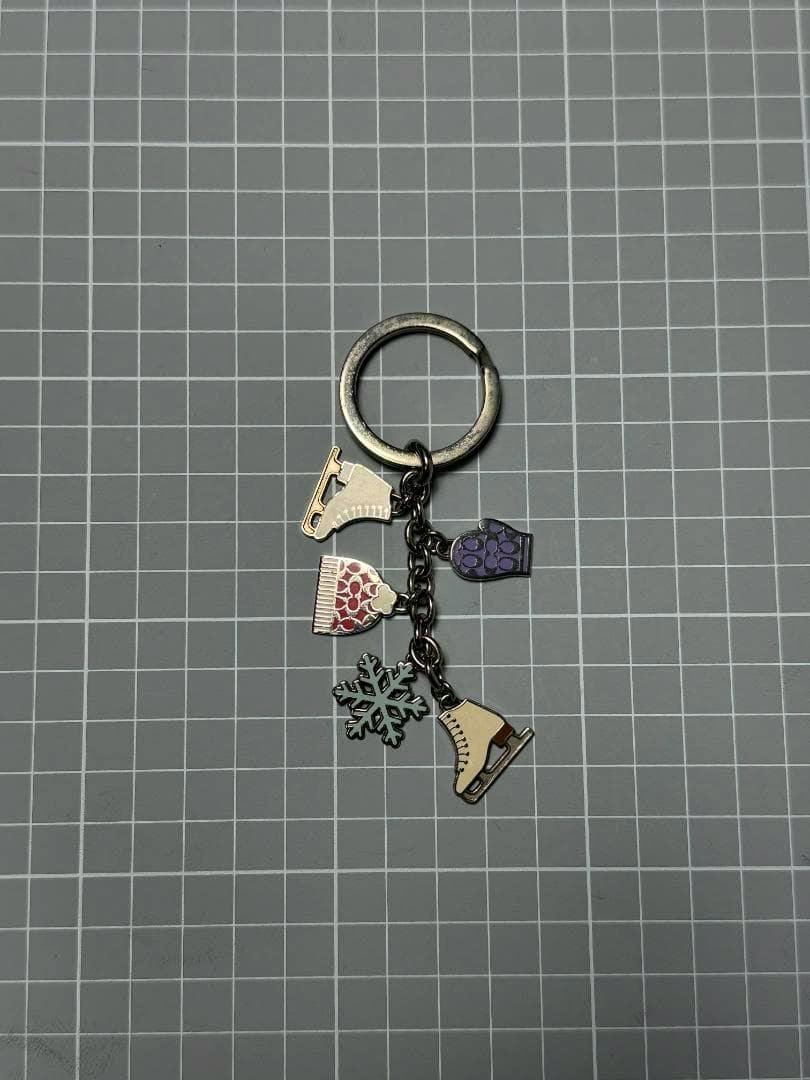 COACH skate-motif bag charm/keychain with ice skate, snowflake, hat, and mitten charms on a metal ring placed on a gray grid background.