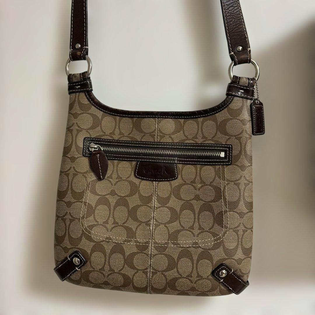 Front view of a brown Coach canvas shoulder bag with signature 'C' pattern, front zip pocket, leather trim, and shoulder strap.