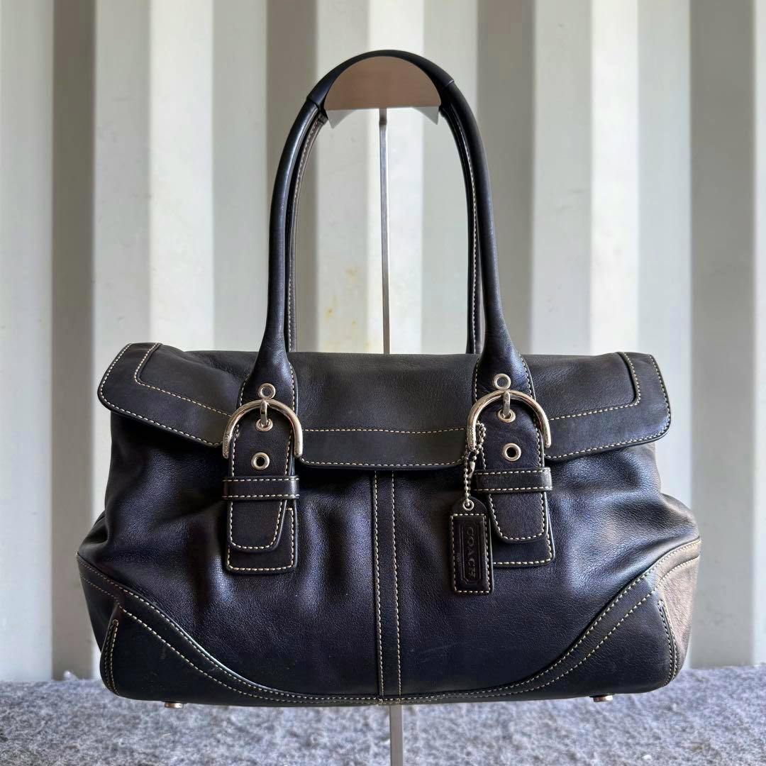 Black Coach Soho leather flap satchel bag with top handles and a hanging tag, shown on a stand.