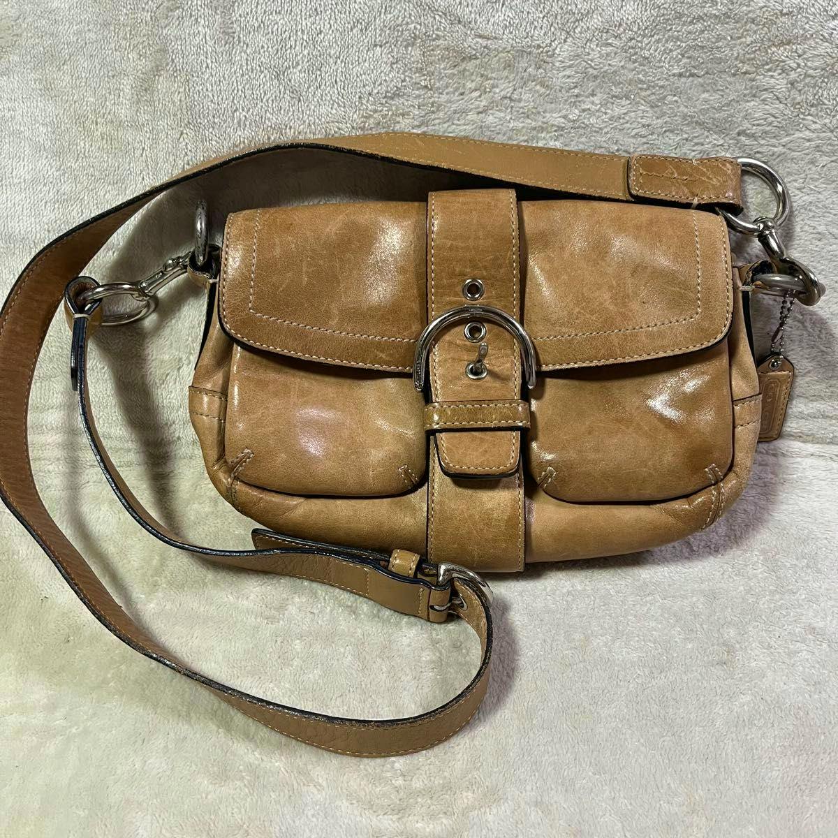 Tan leather COACH Soho shoulder bag with front buckle, two front pockets, strap, and Coach hangtag on a light fabric background.