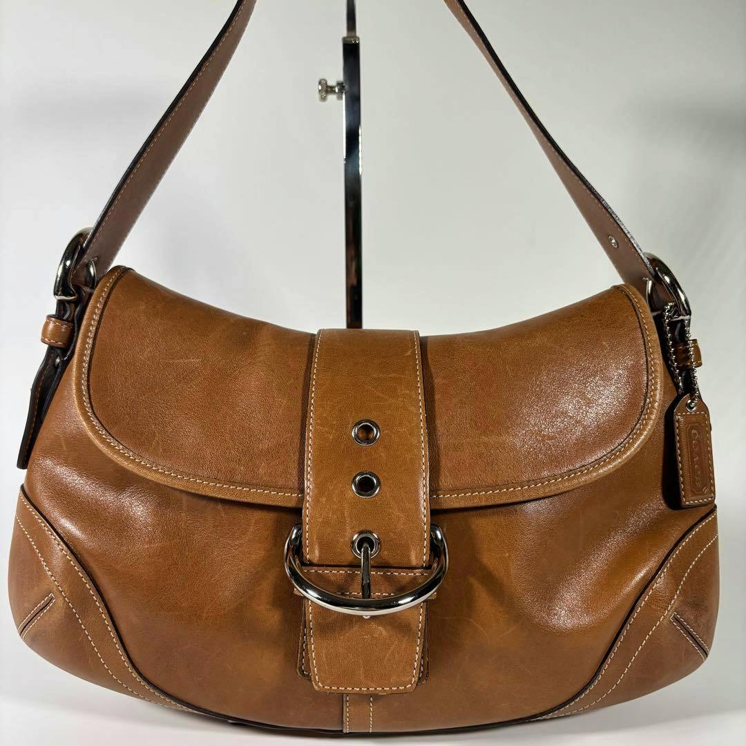 Brown Coach Soho leather shoulder bag with front buckle strap and attached leather tag on a white background.