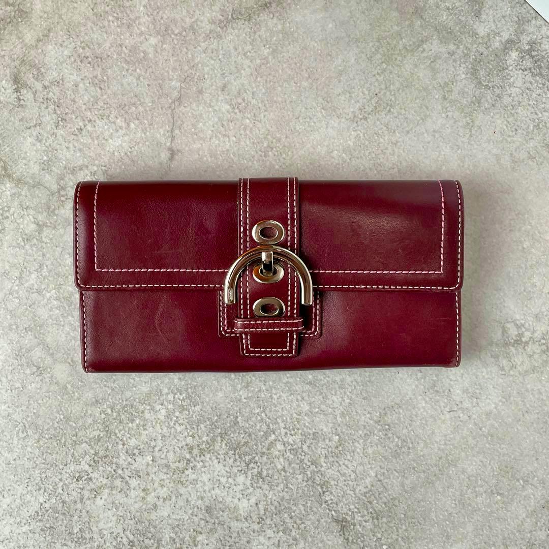 Front of the Coach Soho long cow leather wallet in dark red showing white contrast stitching and a gold-tone buckle closure on a light textured background.