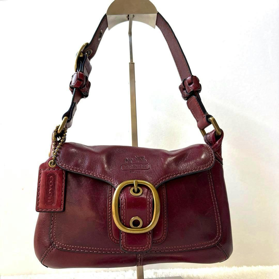 Burgundy Coach one-shoulder leather Soho bag with front brass buckle and tag displayed on a stand.