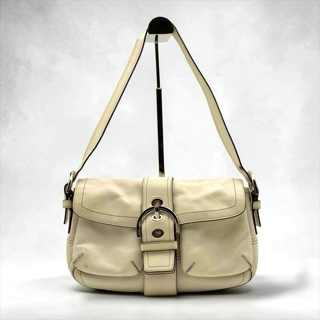 White Coach Soho shoulder bag hanging from a display stand, showing the front flap, buckle, and shoulder strap.