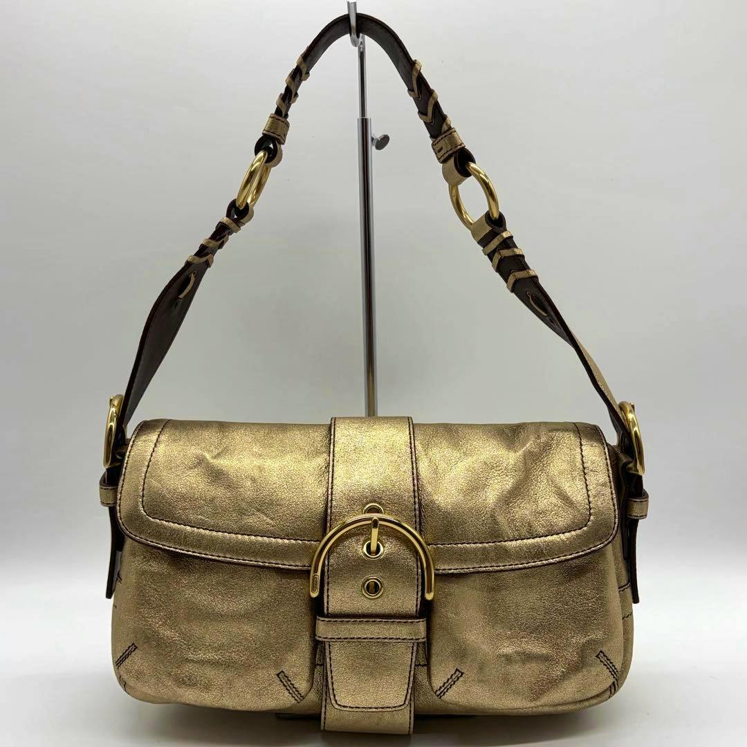 Gold Coach Soho shoulder bag with front buckle and short shoulder strap displayed on a metal stand.