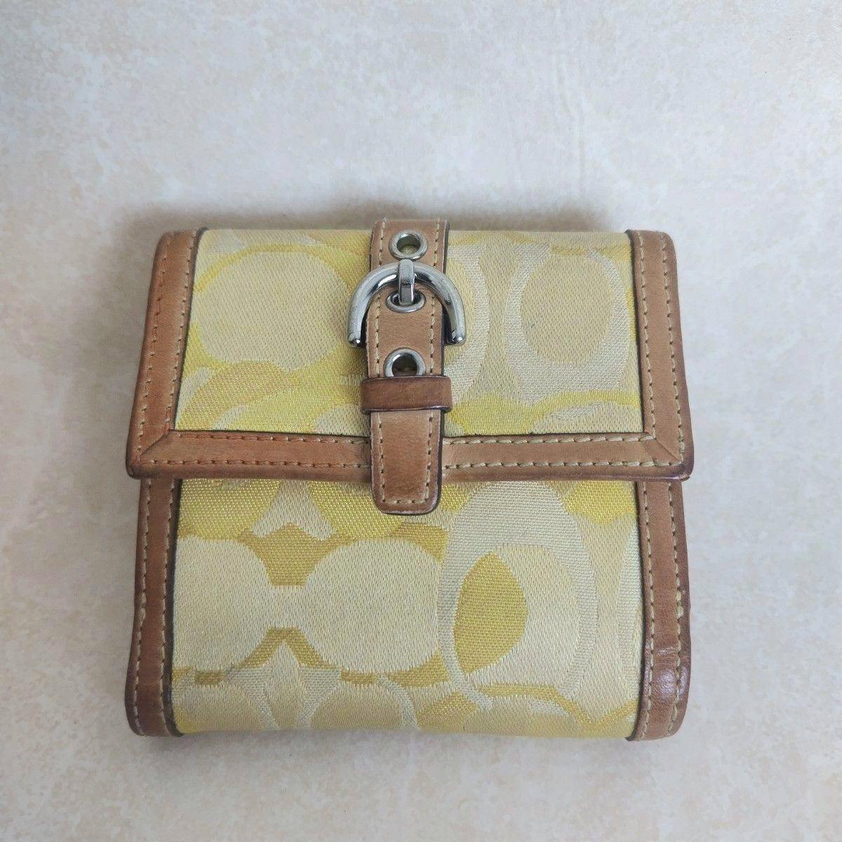 Front view of a yellow Coach signature canvas folding wallet with tan leather trim and a silver buckle closure.