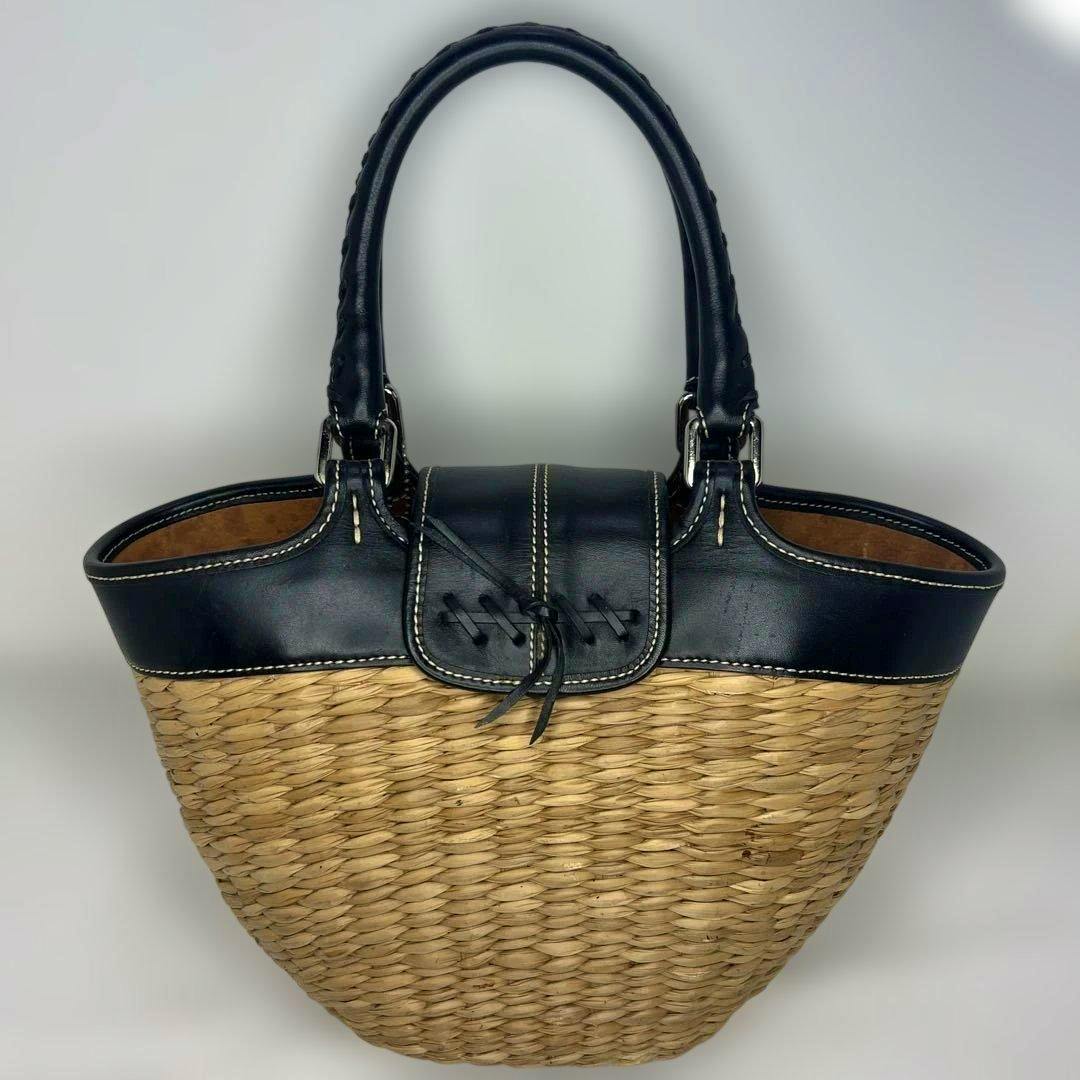 Front view of the straw basket bag showing a woven body with black leather trim, flap, buckle detail, and double handles.