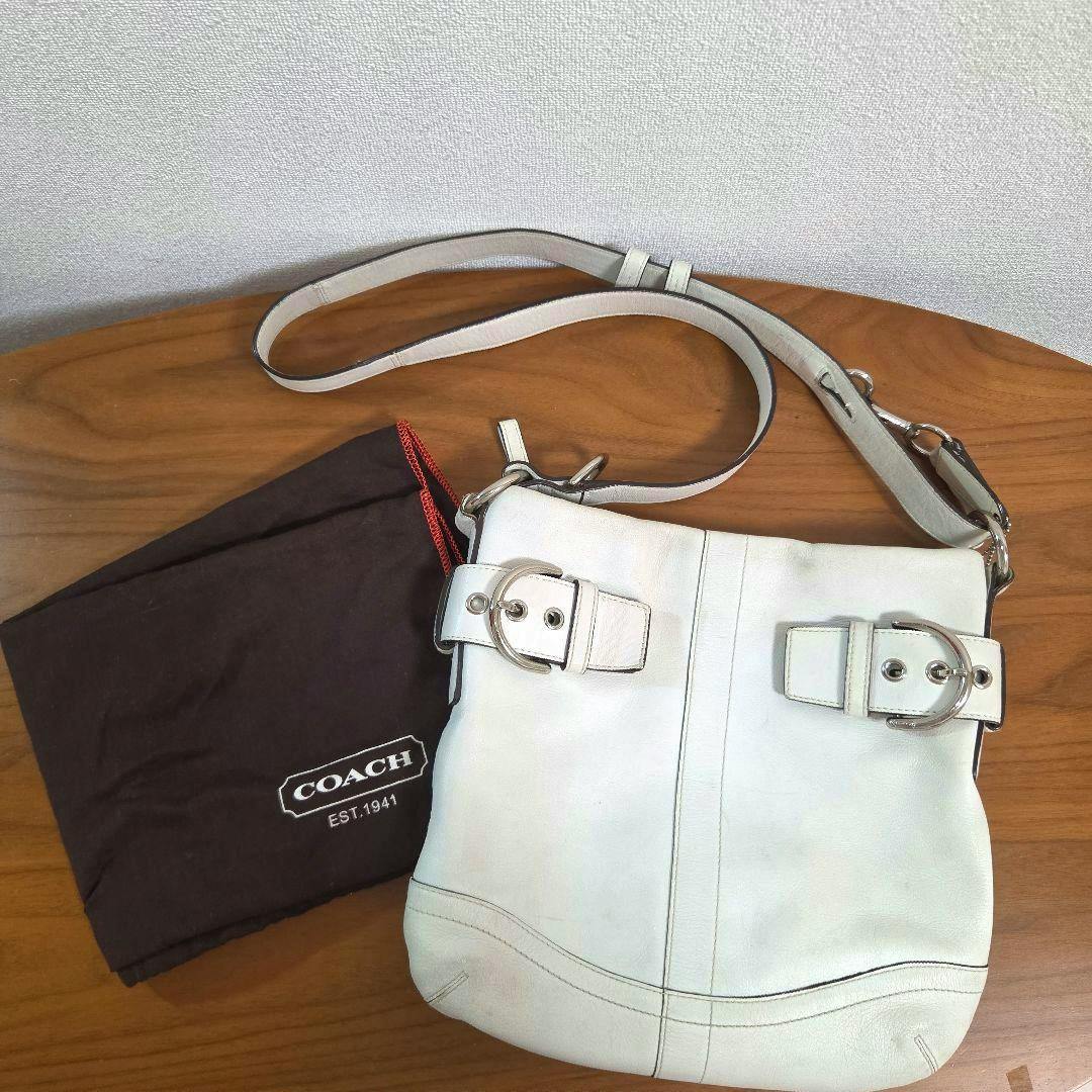 White COACH Soho leather 2WAY shoulder bag lying on a wooden surface next to a brown COACH dust bag.
