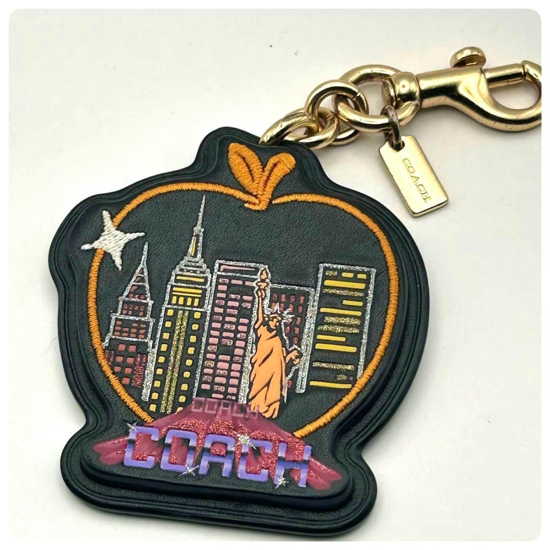 Black apple-shaped leather bag charm with an embroidered city skyline, orange embroidered outline and 'COACH' text, attached to a gold clasp and small Coach tag.