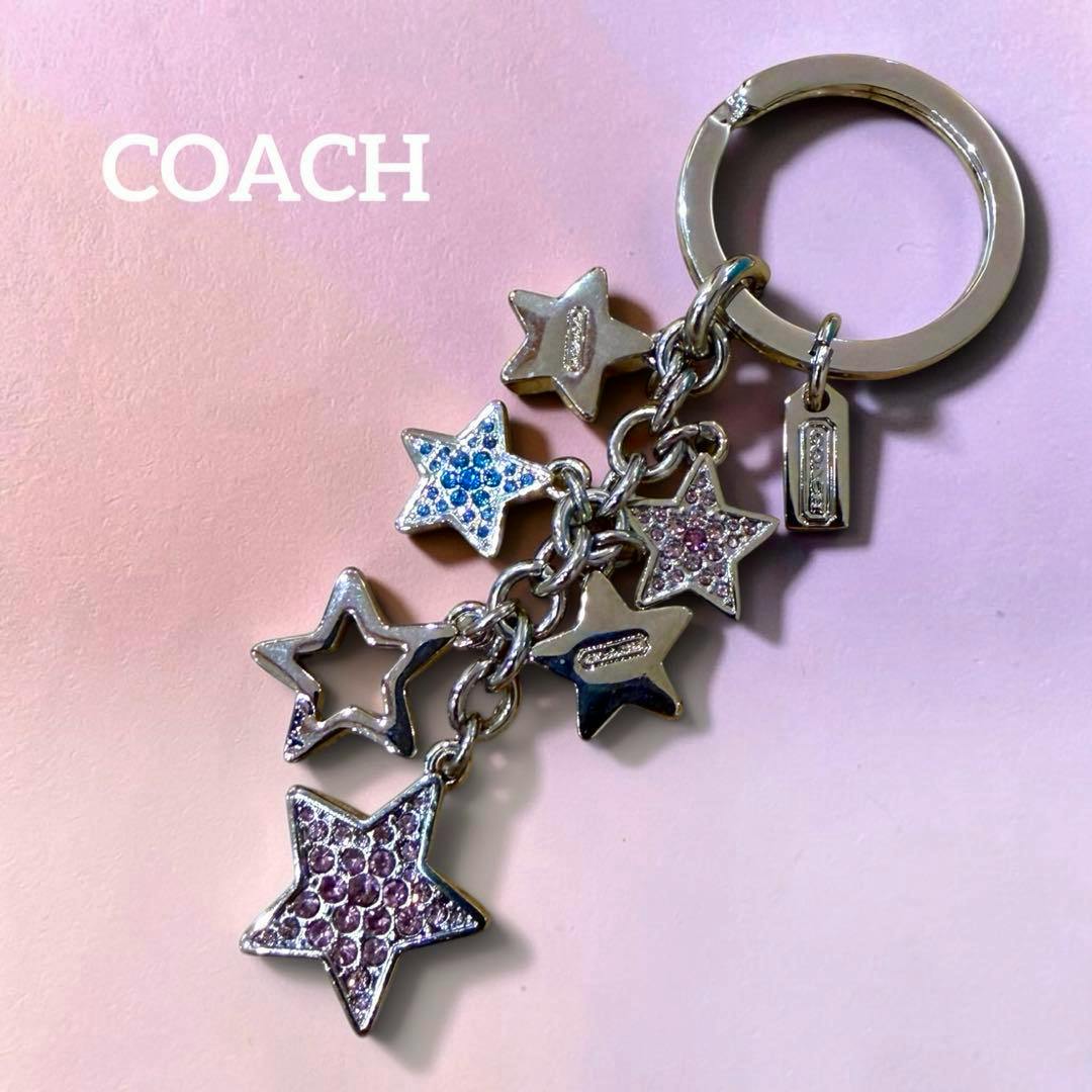 Silver Coach star bag charm with multiple star pendants, some set with pink and blue crystals, shown on a pink background with COACH text.
