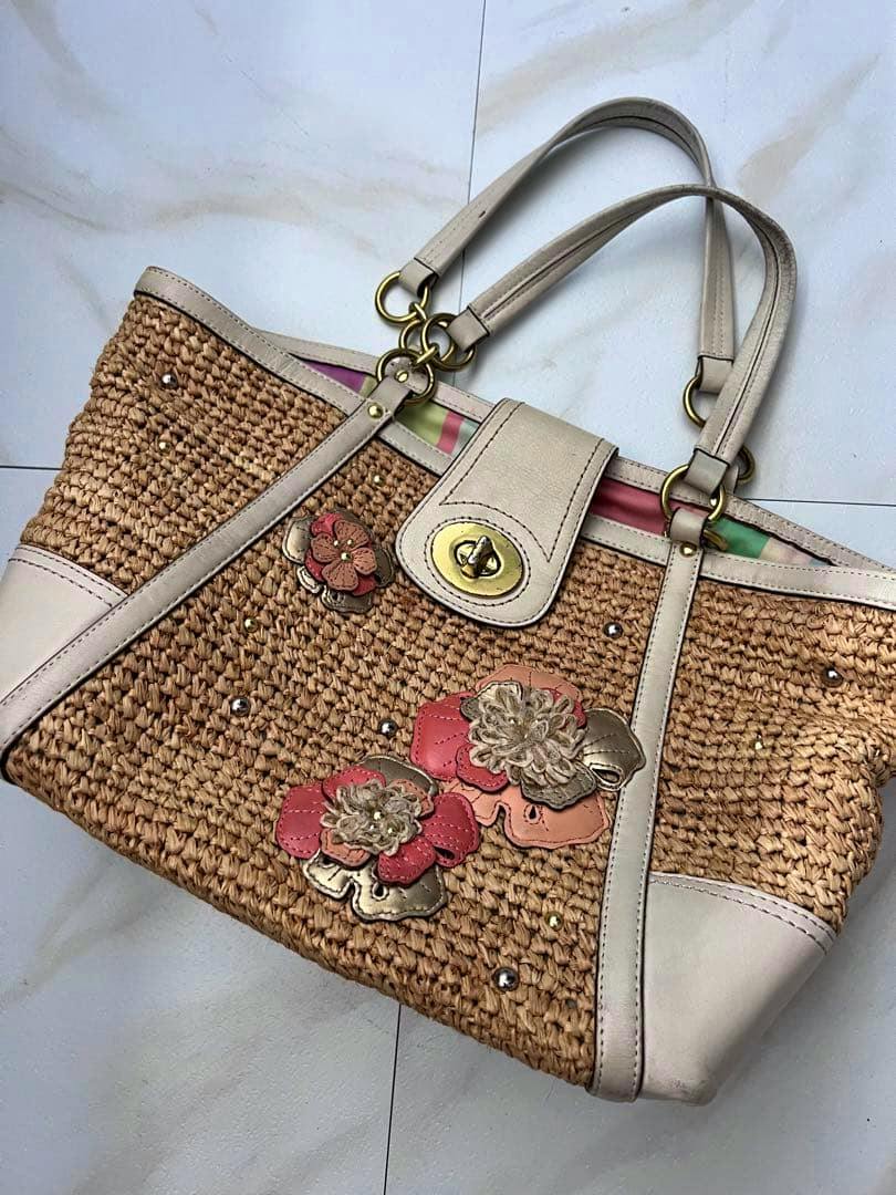 Coach straw tote bag with light leather trim, floral appliqués, and a gold turn-lock clasp lying on a tiled floor.