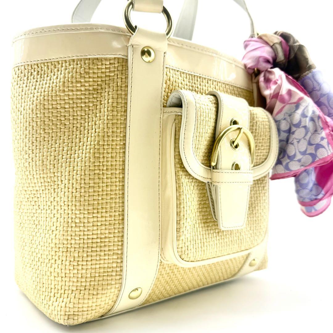 Close-up of a straw Coach handbag showing light leather trim, gold hardware, and a pink patterned scarf tied to the handle.