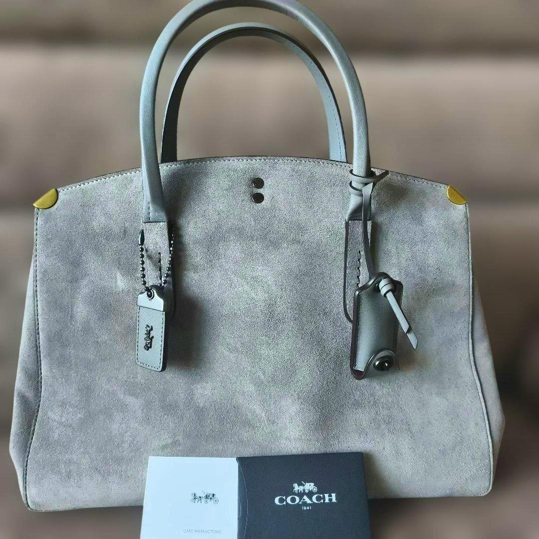 Gray COACH suede handbag with double top handles, leather tags, and gold corner protectors shown on a neutral background with COACH booklets at the front.