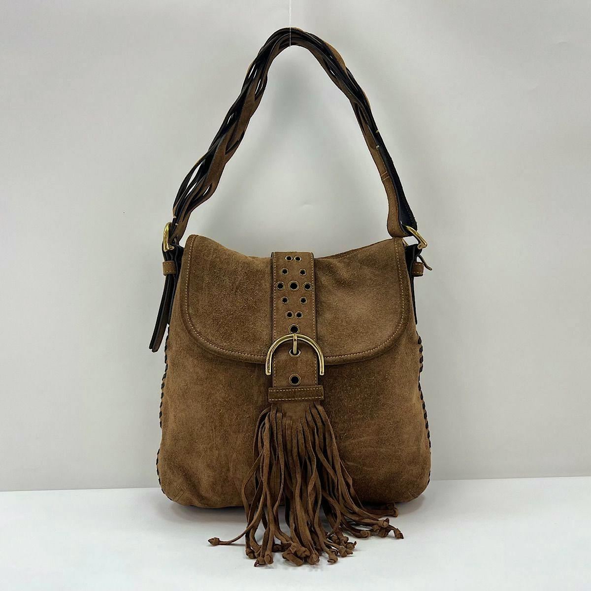 Front view of a brown COACH suede leather shoulder bag with belt-style buckle and long fringe.