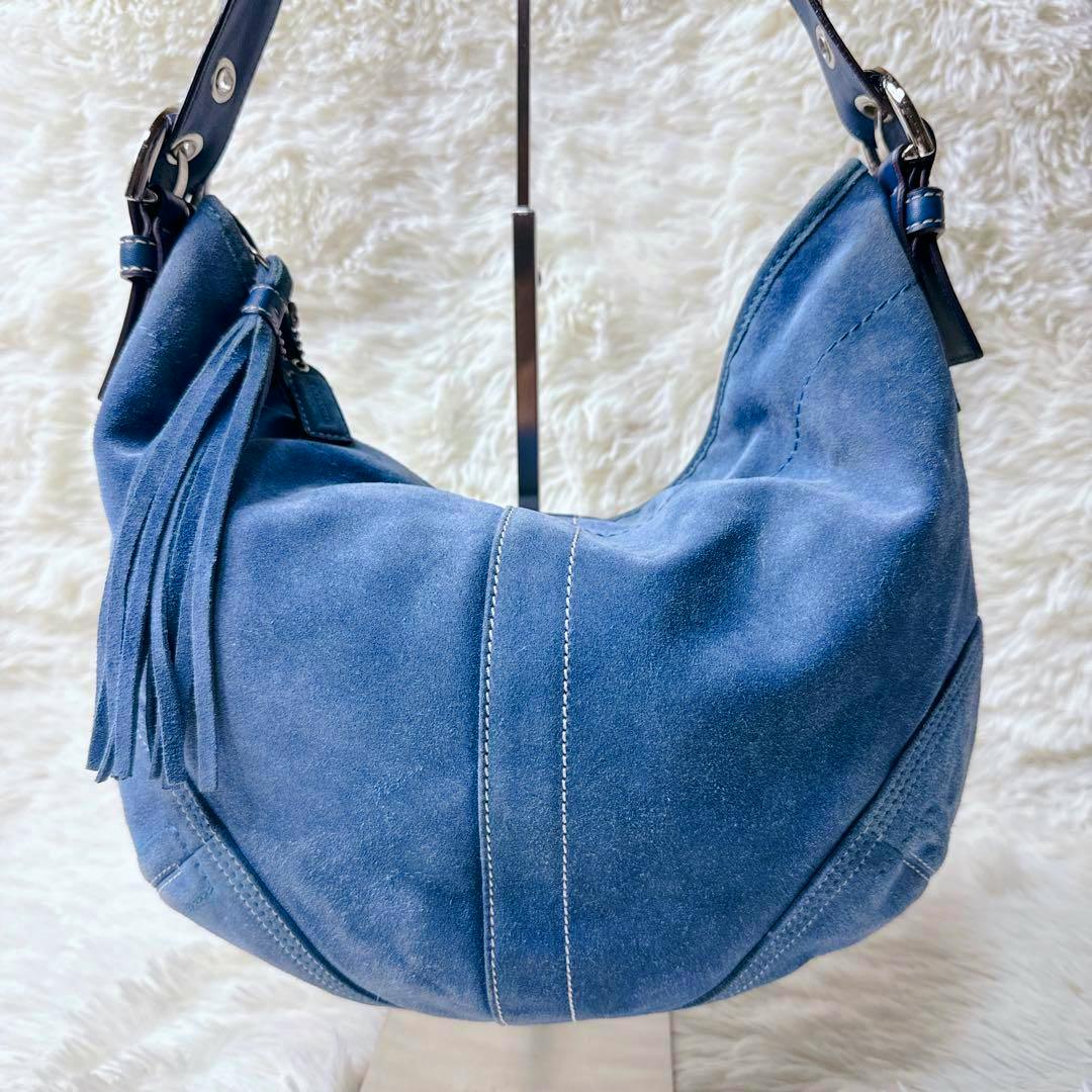 Blue suede COACH shoulder bag hanging on a stand against a white fur background.