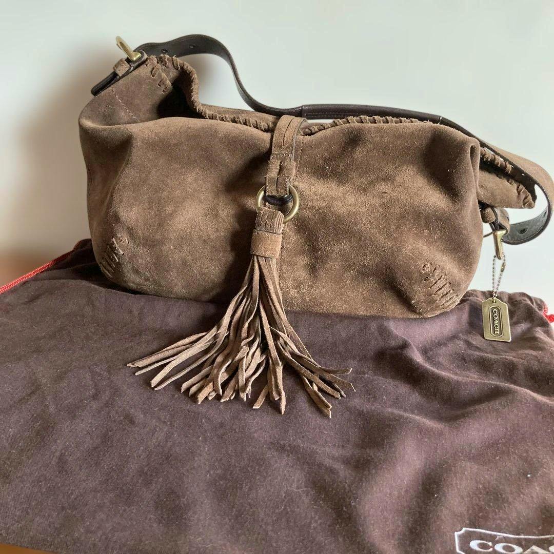 Brown suede Coach shoulder bag with a front fringe tassel resting on a brown Coach dust bag.