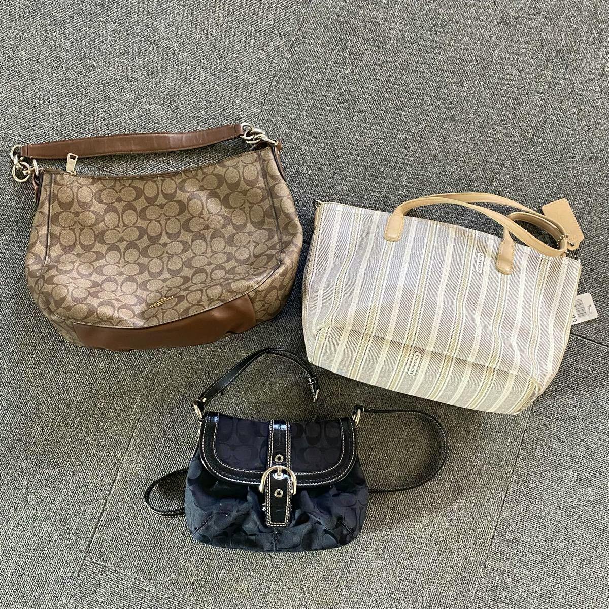 Three Coach bags laid on a gray carpet: a brown monogram shoulder bag, a light striped tote, and a small black bag.