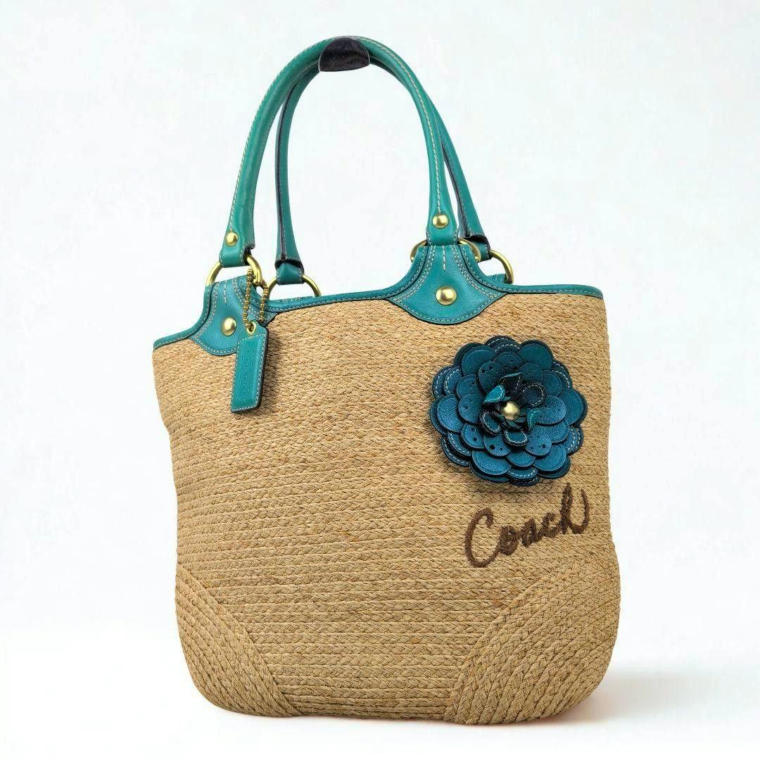 Front view of a Coach woven straw tote bag with teal leather handles, a teal layered flower applique, and "Coach" embroidered script.