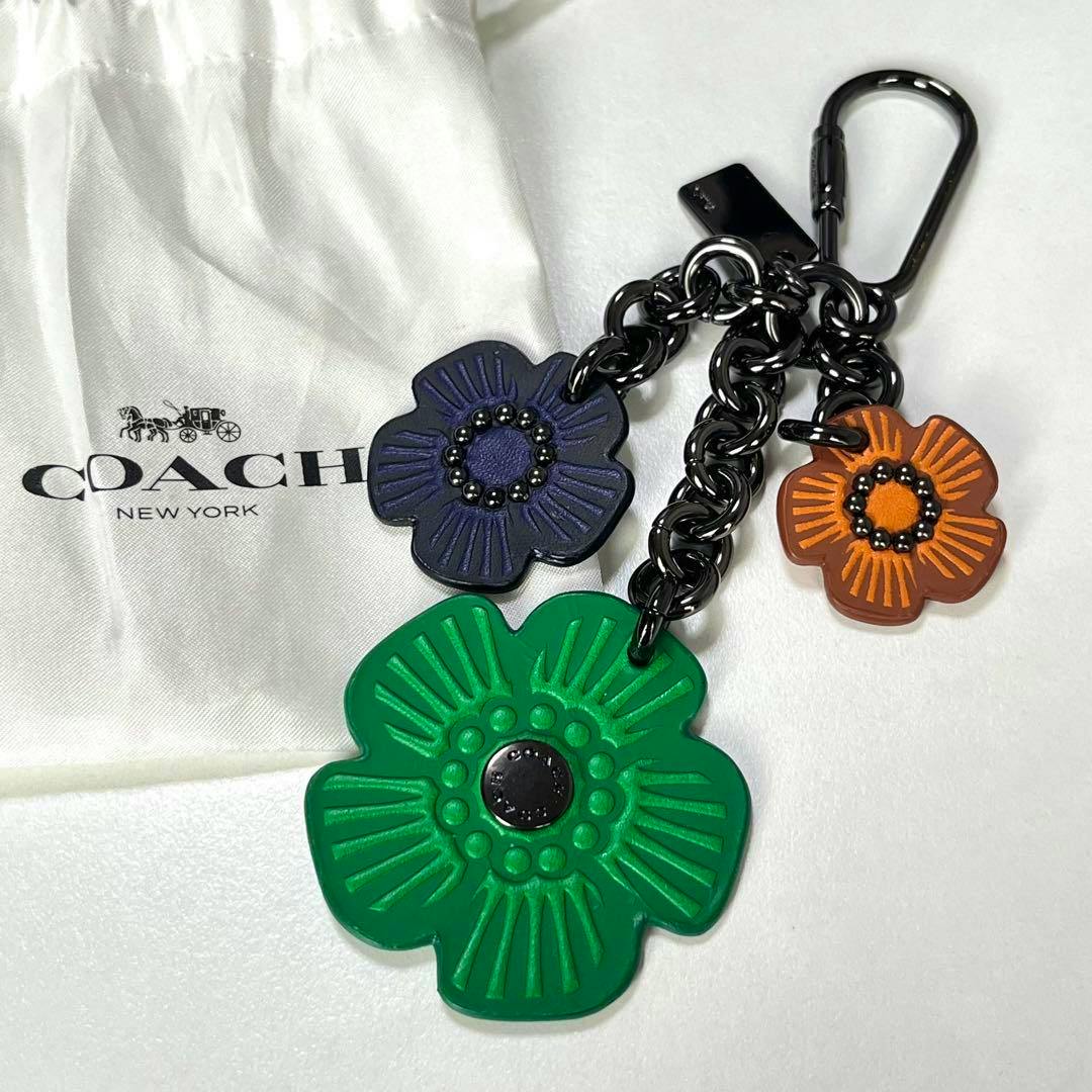 COACH Tea Rose flower keychain bag charm with three colorful flower pendants on a black chain, displayed on a white Coach dust bag.