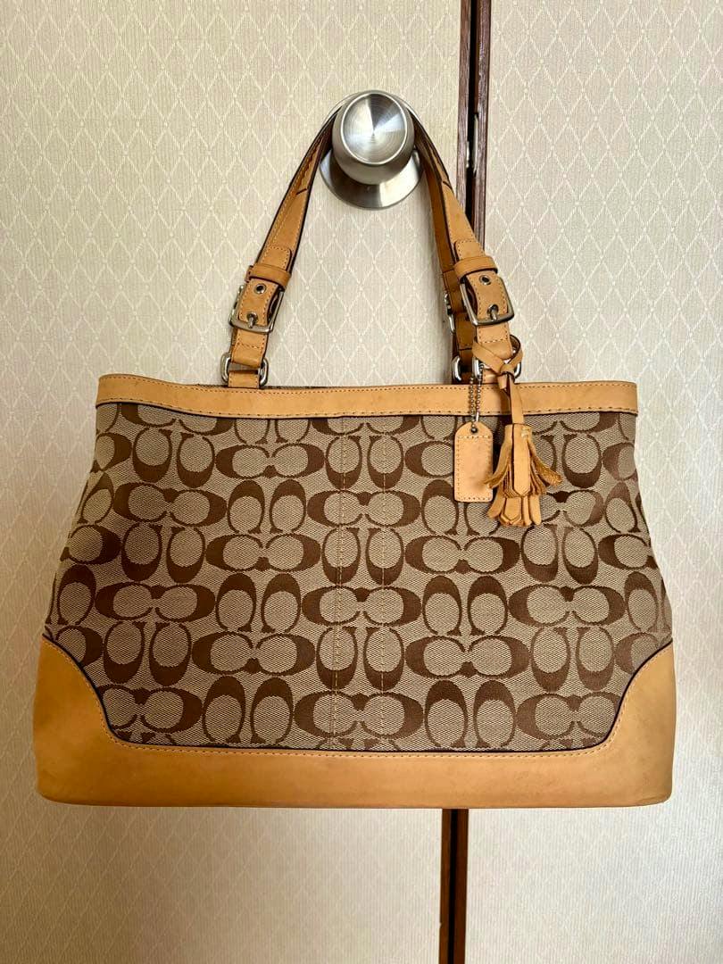 Brown and beige COACH canvas tote bag with repeating logo pattern, leather trim, and a tassel hangtag hanging from a door knob.