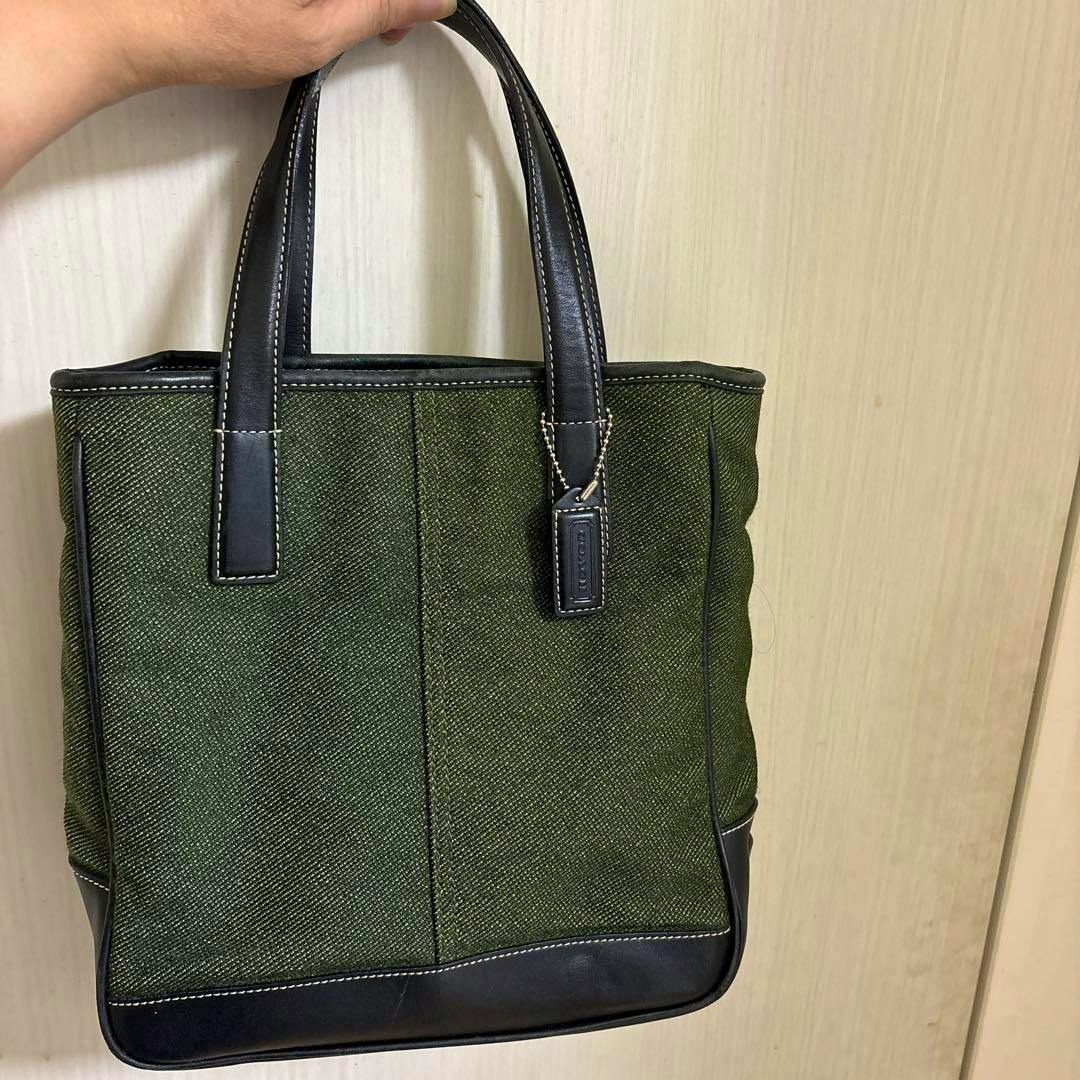 Green COACH tote bag with black leather handles and trim held by a hand against a light background.