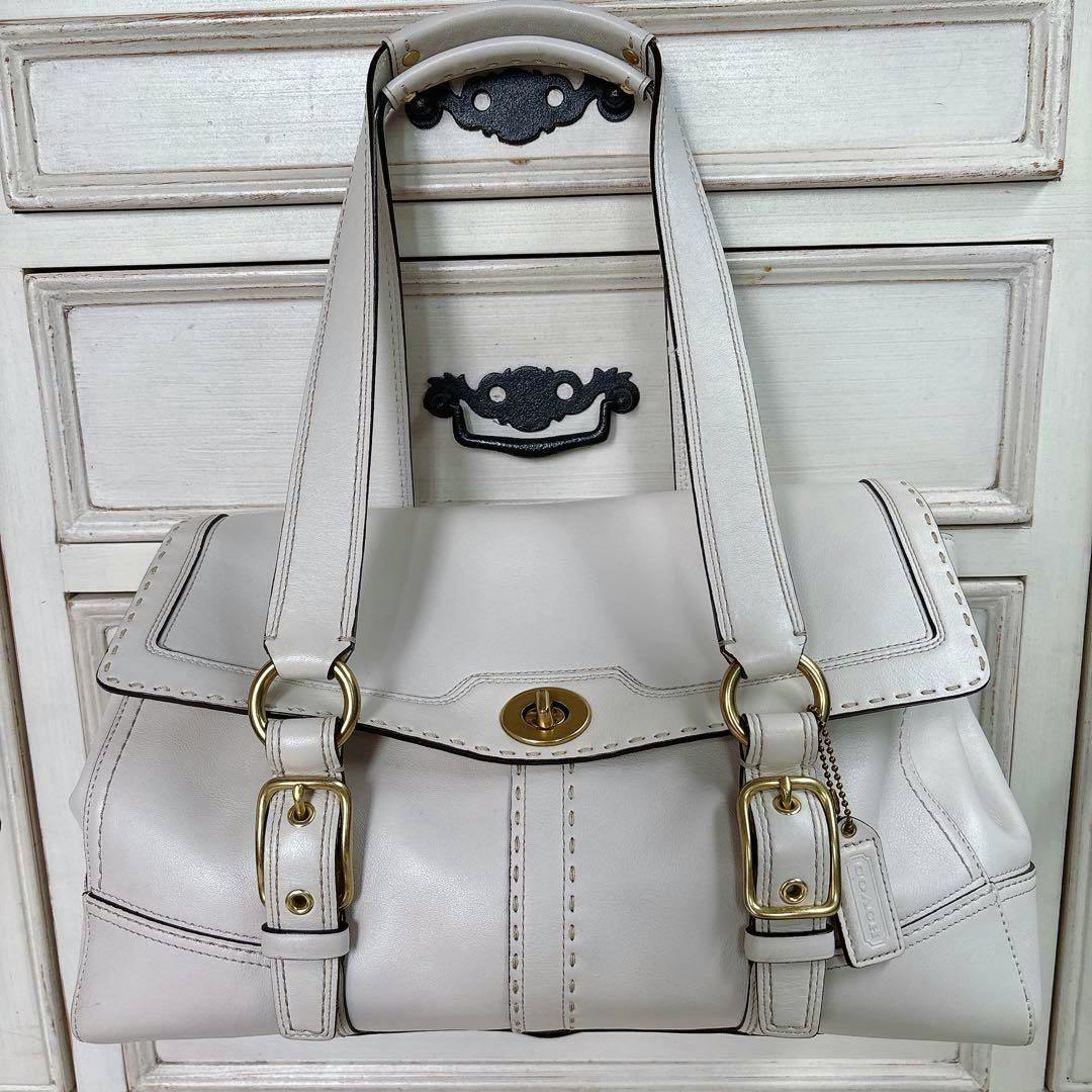 White COACH turn-lock leather shoulder tote bag with gold hardware hanging on a painted wooden cabinet.