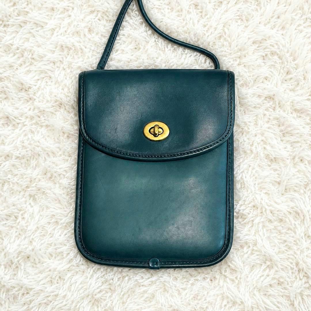 Front view of the blue-green COACH cow leather smartphone shoulder bag with a turn-lock closure on a white textured surface.
